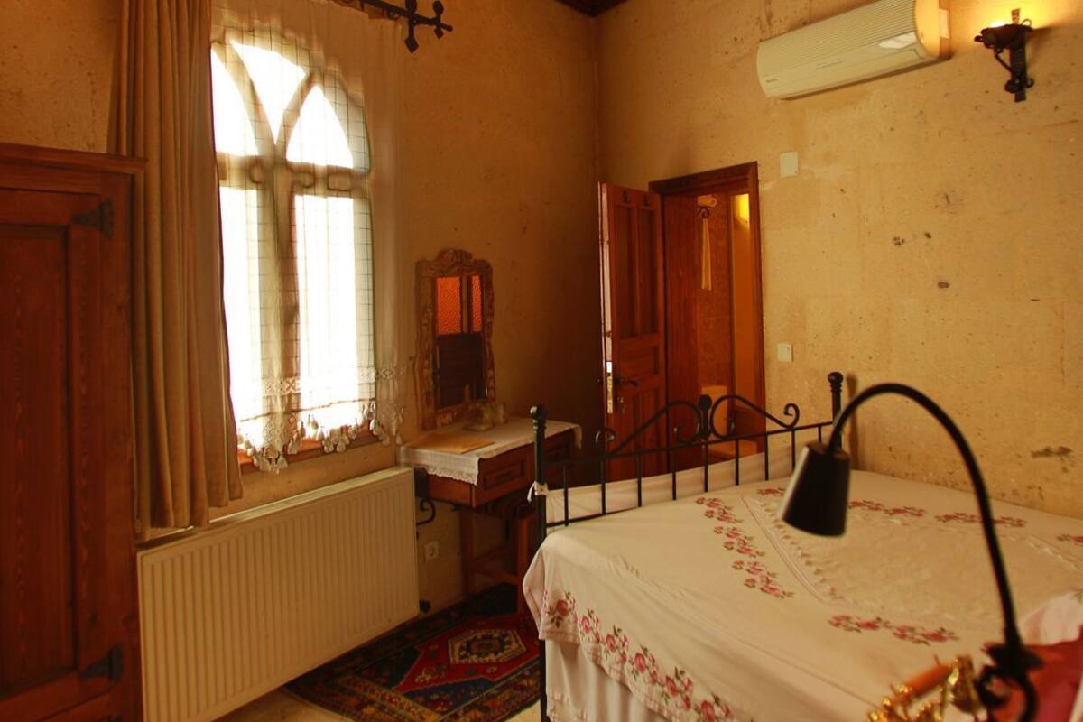 A bedroom with a wooden ceiling, a bed with a floral bedspread, a radiator, a window with curtains, and a wall-mounted light fixture.