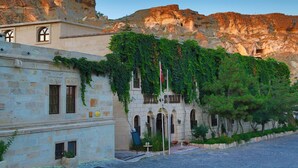Un edificio con paredes de piedra y ventanas arqueadas, rodeado de vegetación y con una montaña rocosa de fondo.