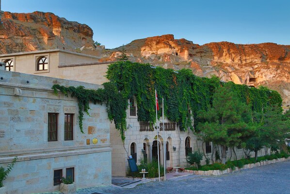 A building with stone walls and arched windows, surrounded by greenery and a rocky mountain backdrop.