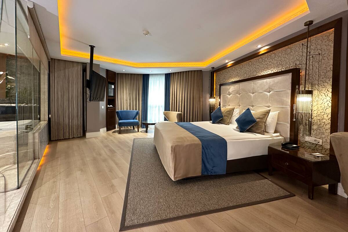 Una habitación de hotel moderna con una cama grande, una zona de estar con un sillón azul, un separador de vidrio y un ventanal con cortinas.