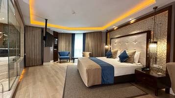 A modern hotel room with a large bed, a sitting area with a blue armchair, a glass partition, and a large window with curtains.