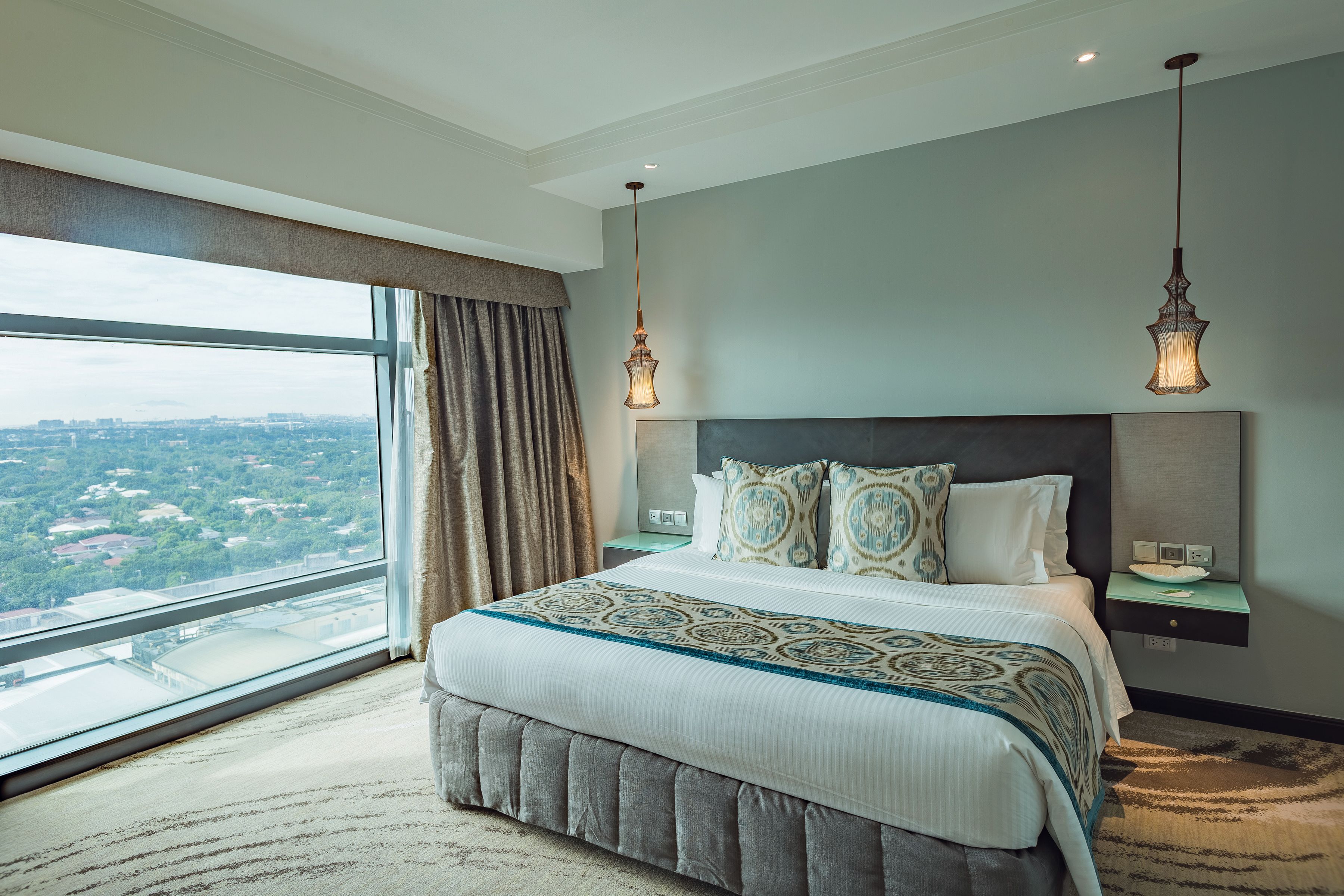 Un dormitorio moderno con una cama grande, dos mesitas de noche y vistas a la ciudad, con un paisaje urbano visible a través de la ventana.