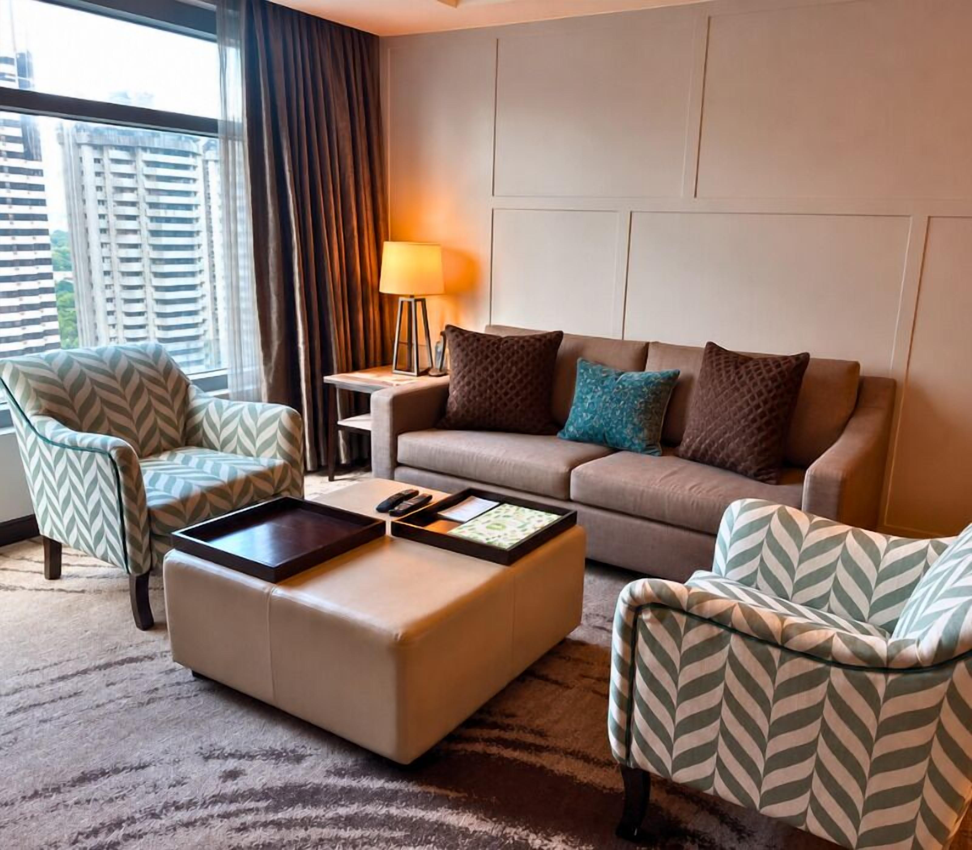 Une chambre d’hôtel avec un canapé, deux fauteuils, une table basse, une lampe et une vue sur la ville.