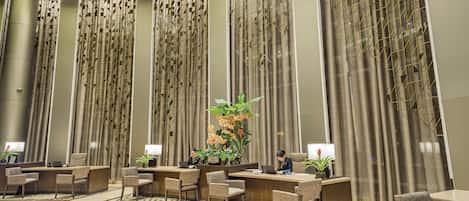 A hotel lobby with a reception desk, several chairs, a large decorative plant, and tall curtains with a geometric pattern.