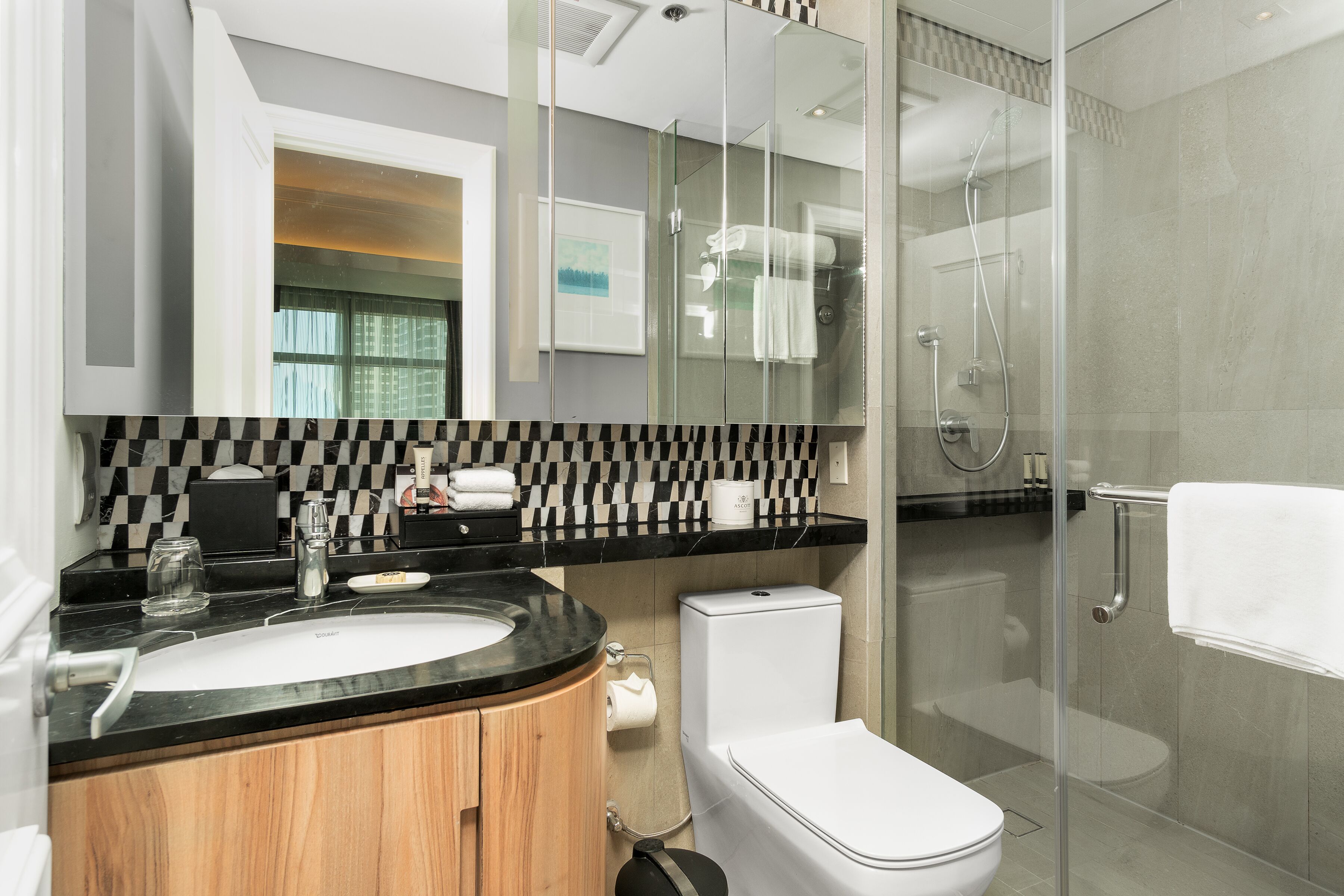 A modern bathroom with a glass-enclosed shower, a wooden vanity, a white toilet, and a black and white checkered backsplash.