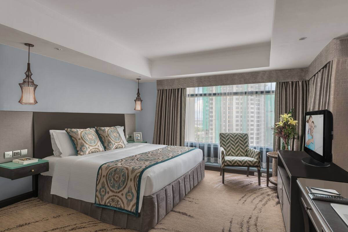Habitación de hotel con una cama grande, un escritorio, una silla, un televisor y una ventana con vistas a la ciudad.