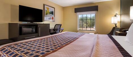A hotel room with a large bed, a flat-screen TV, a microwave, a desk, and a window with a view of trees.