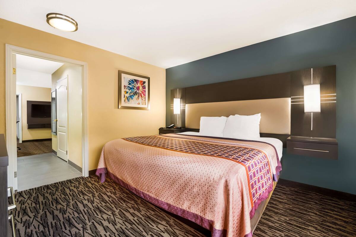 A hotel room with a large bed, a headboard with wall-mounted lights, a carpeted floor, and a door leading to a hallway.