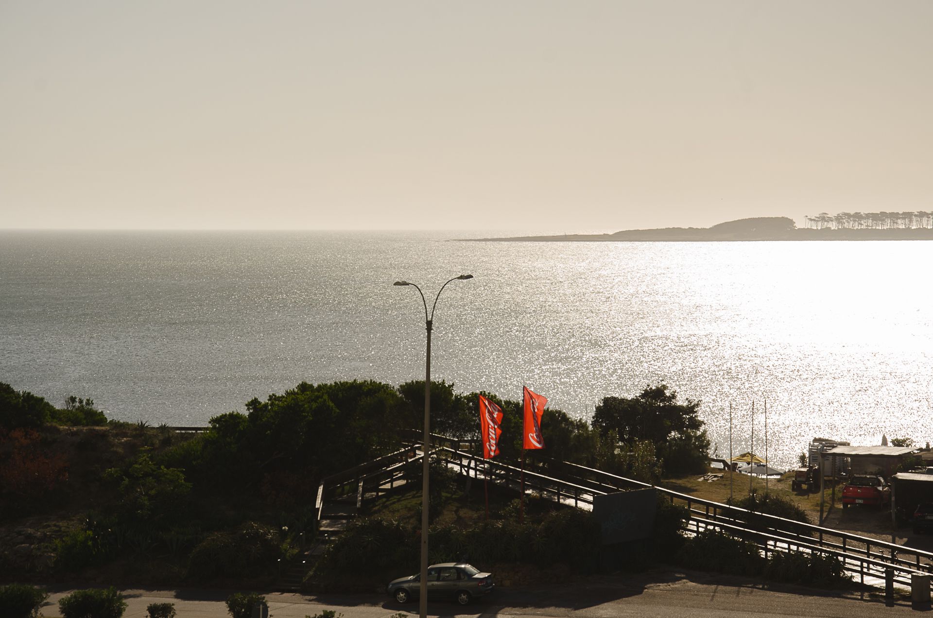 Une route côtière avec un pont menant à une plage, agrémentée de drapeaux orange et offrant une vue sur une côte lointaine.