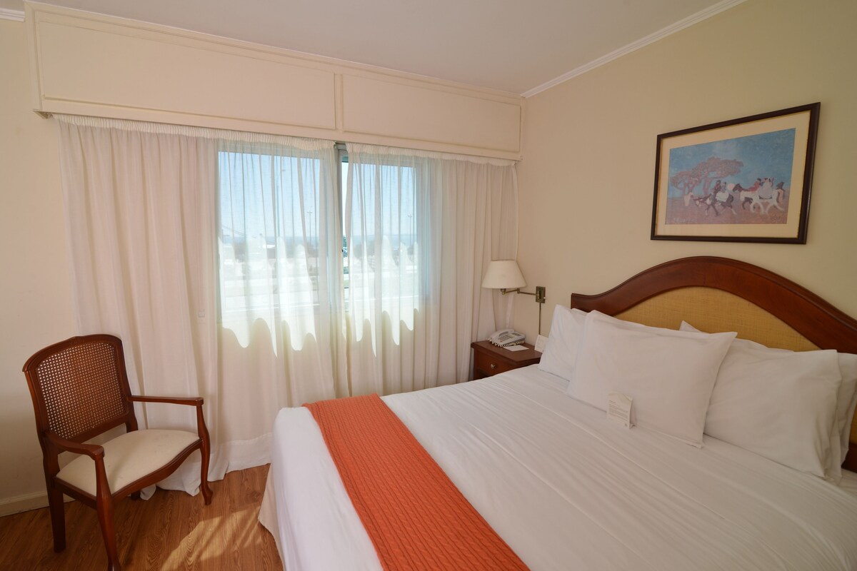 Une chambre d’hôtel avec un grand lit, une chaise en bois, une table de chevet avec une lampe et un tableau au mur.