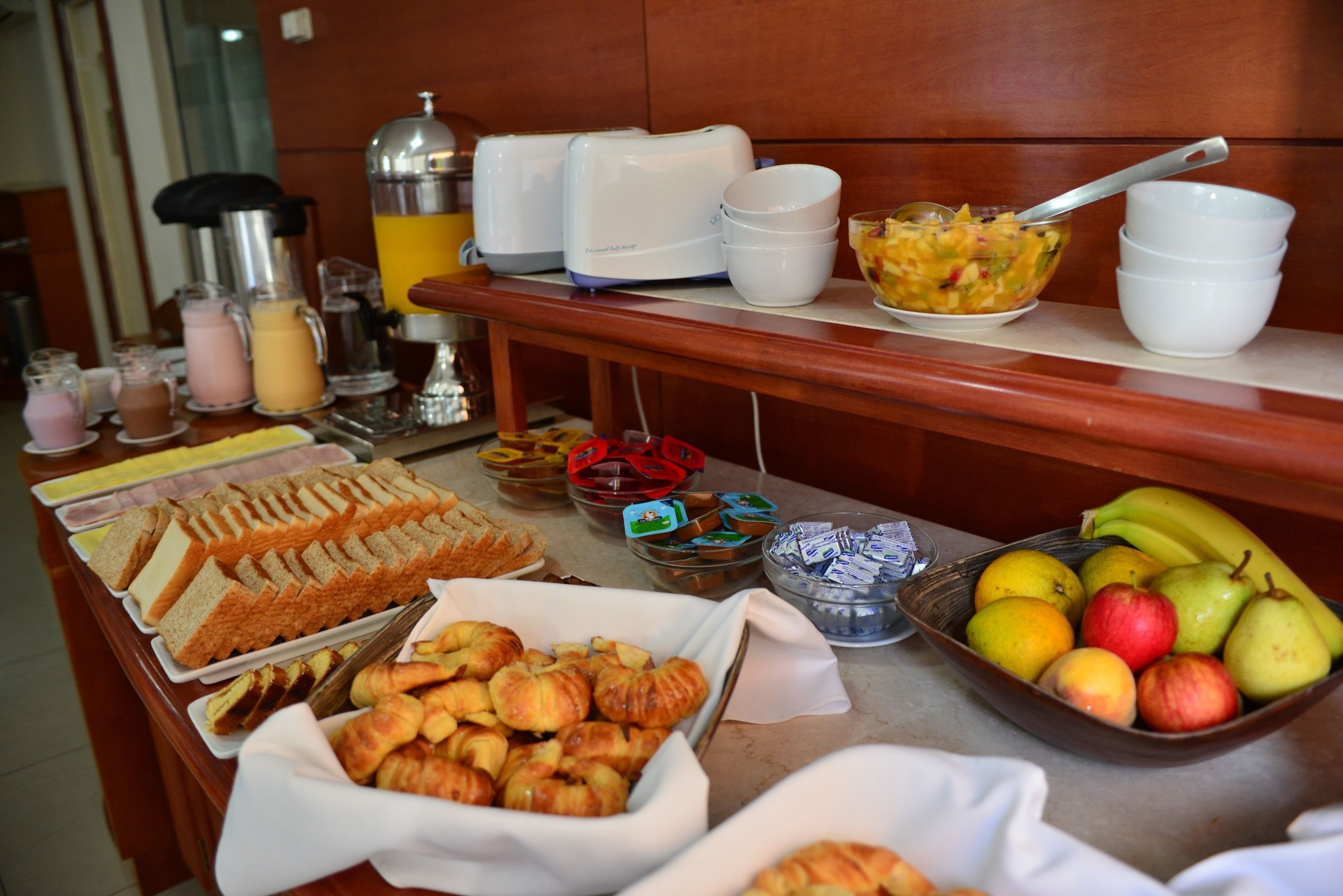 Buffet com diversos alimentos, incluindo pão, pastelaria, frutas e bebidas.