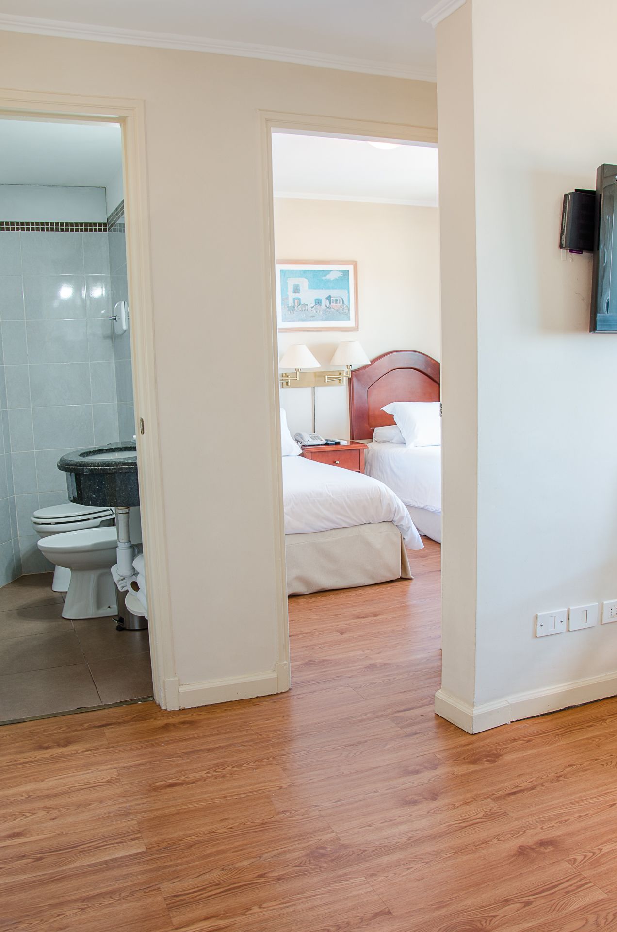 Habitación de hotel con piso de madera, baño con ducha y retrete, y cama con ropa blanca.