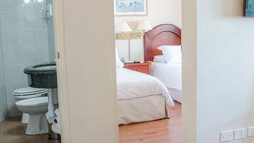 Habitación de hotel con piso de madera, baño con ducha y retrete, y cama con ropa blanca.