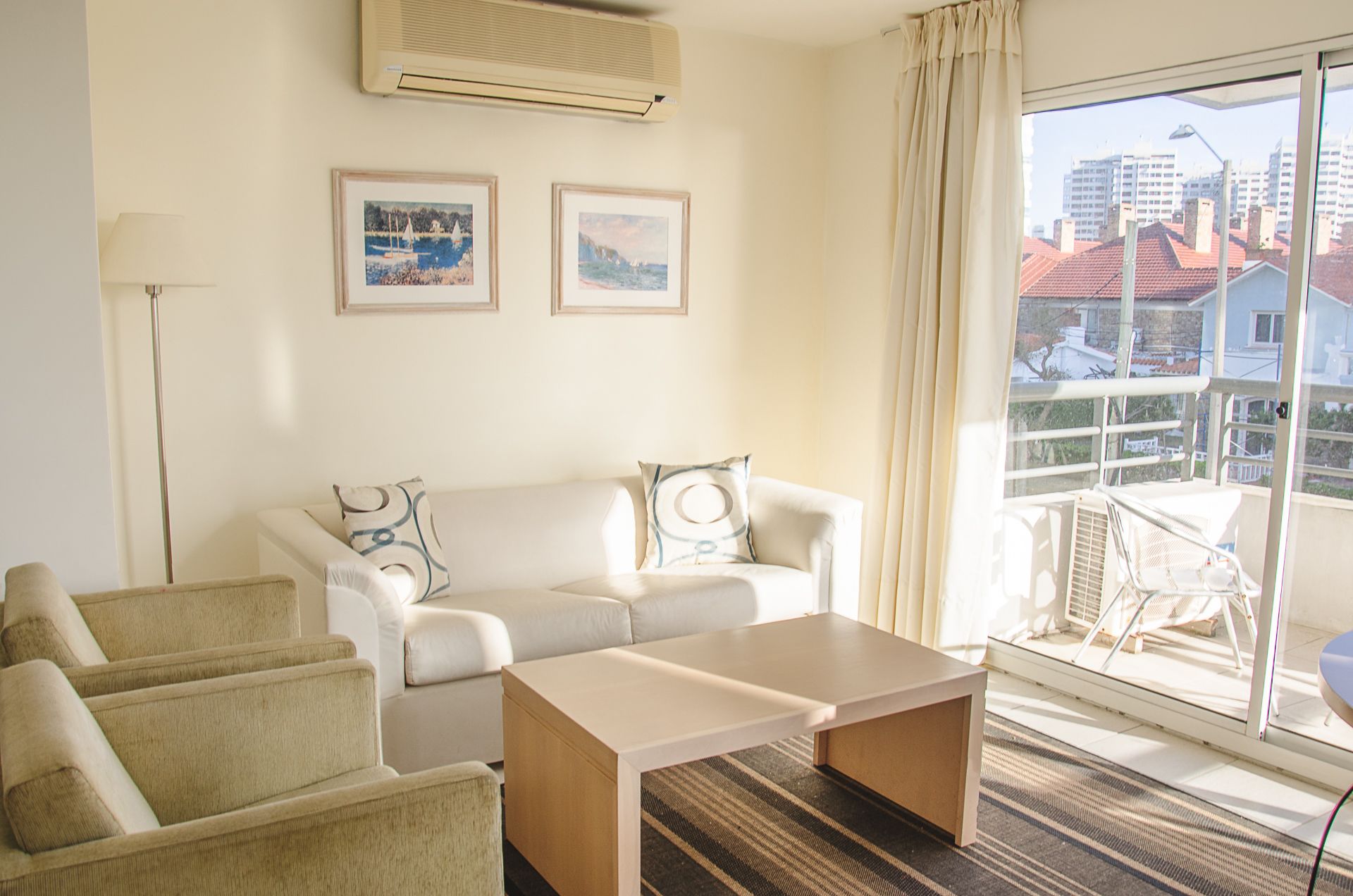 A modern living room with a sofa, armchairs, a coffee table, and a balcony with a view of buildings.