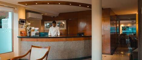 A hotel reception area with a receptionist, a reception desk, a chair, and a view of the restaurant.