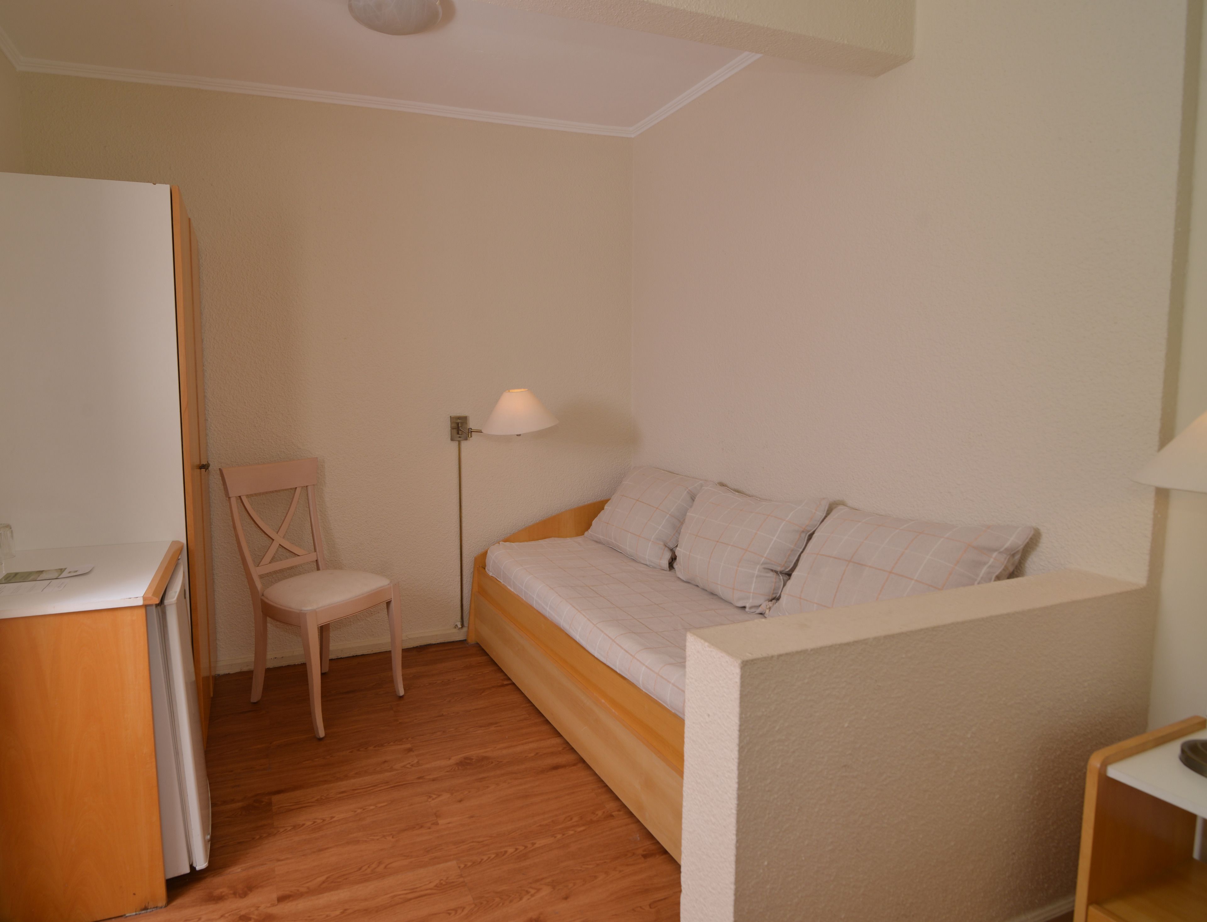Una habitación pequeña y acogedora con piso de madera, una cama con colchón y almohadas blancas, una silla, un escritorio con lámpara y un aplique de pared.