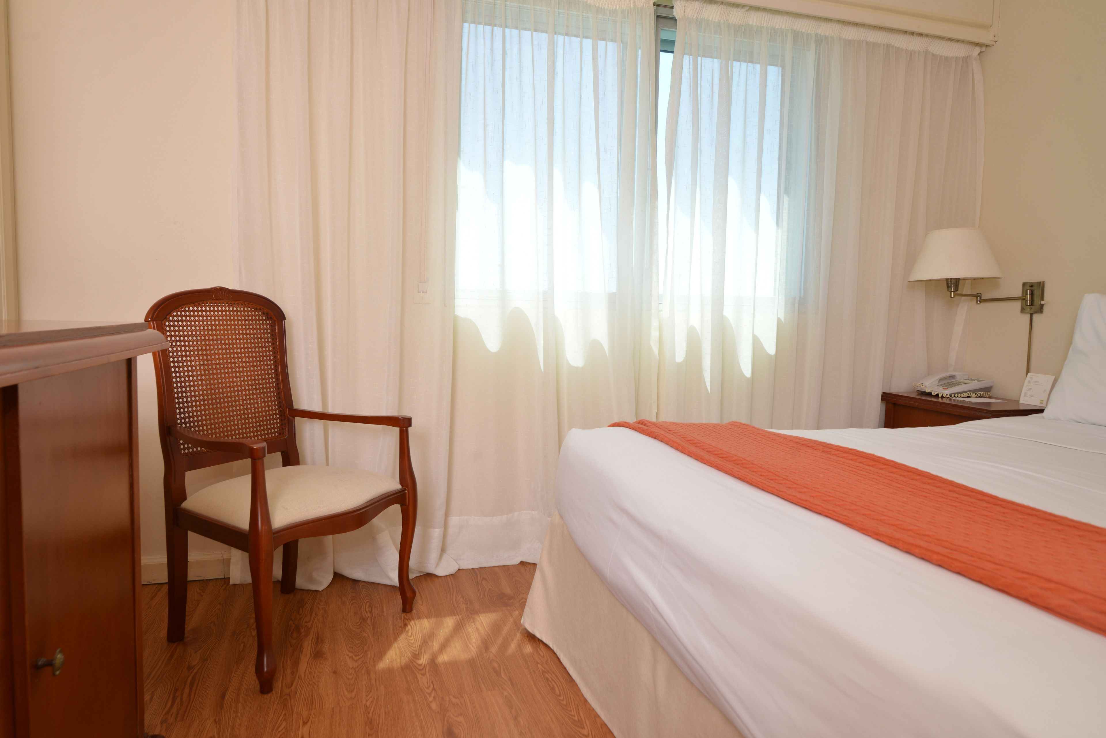 A hotel room with a bed, a chair, a desk, a lamp, and a window with curtains.