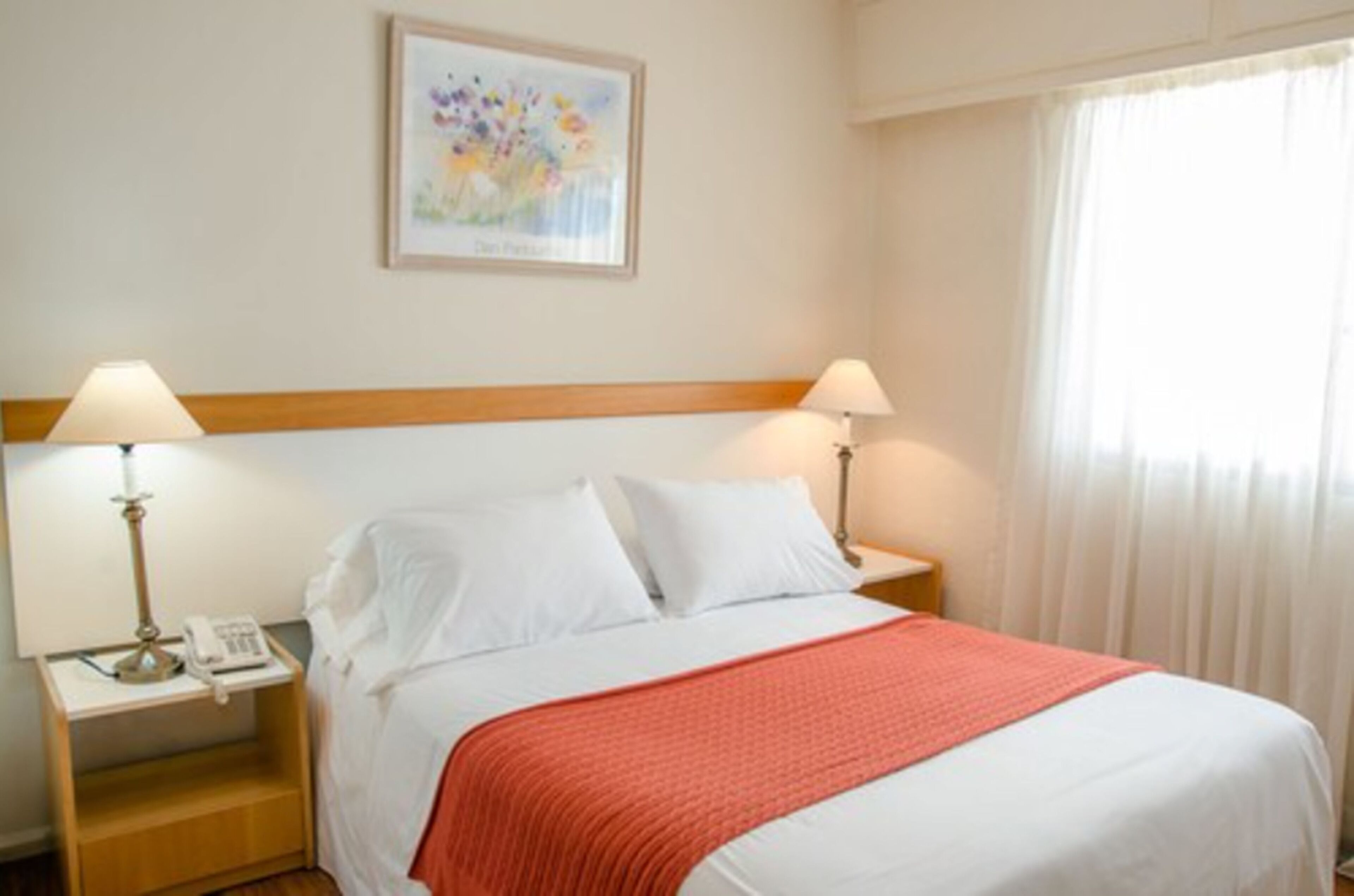 Quarto de hotel com uma cama, duas mesas de cabeceira, uma lâmpada em cada mesa, um telefone em uma das mesas e um quadro na parede.