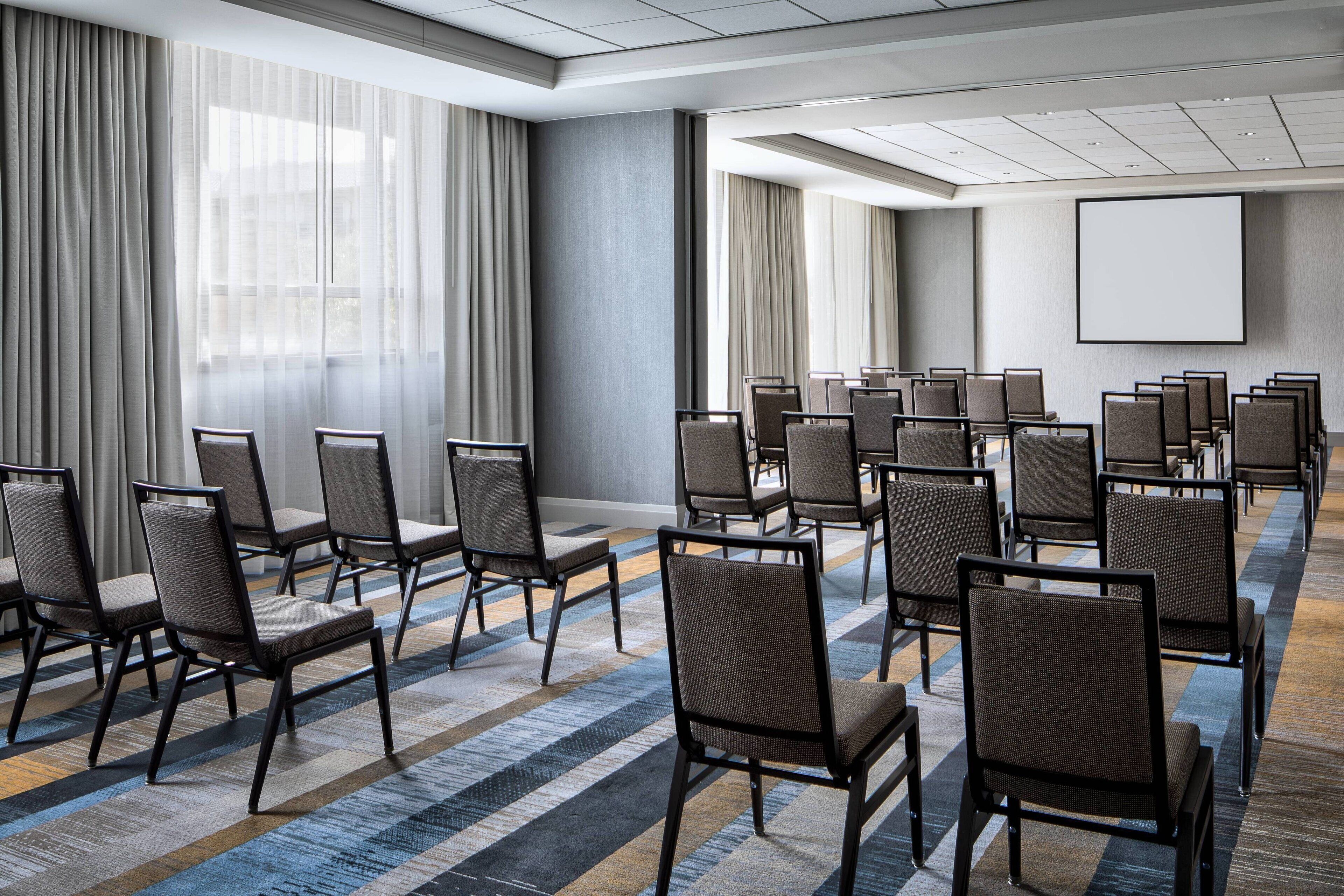 Un salón de conferencias con filas de sillas, una pantalla grande y amplios ventanales con cortinas.