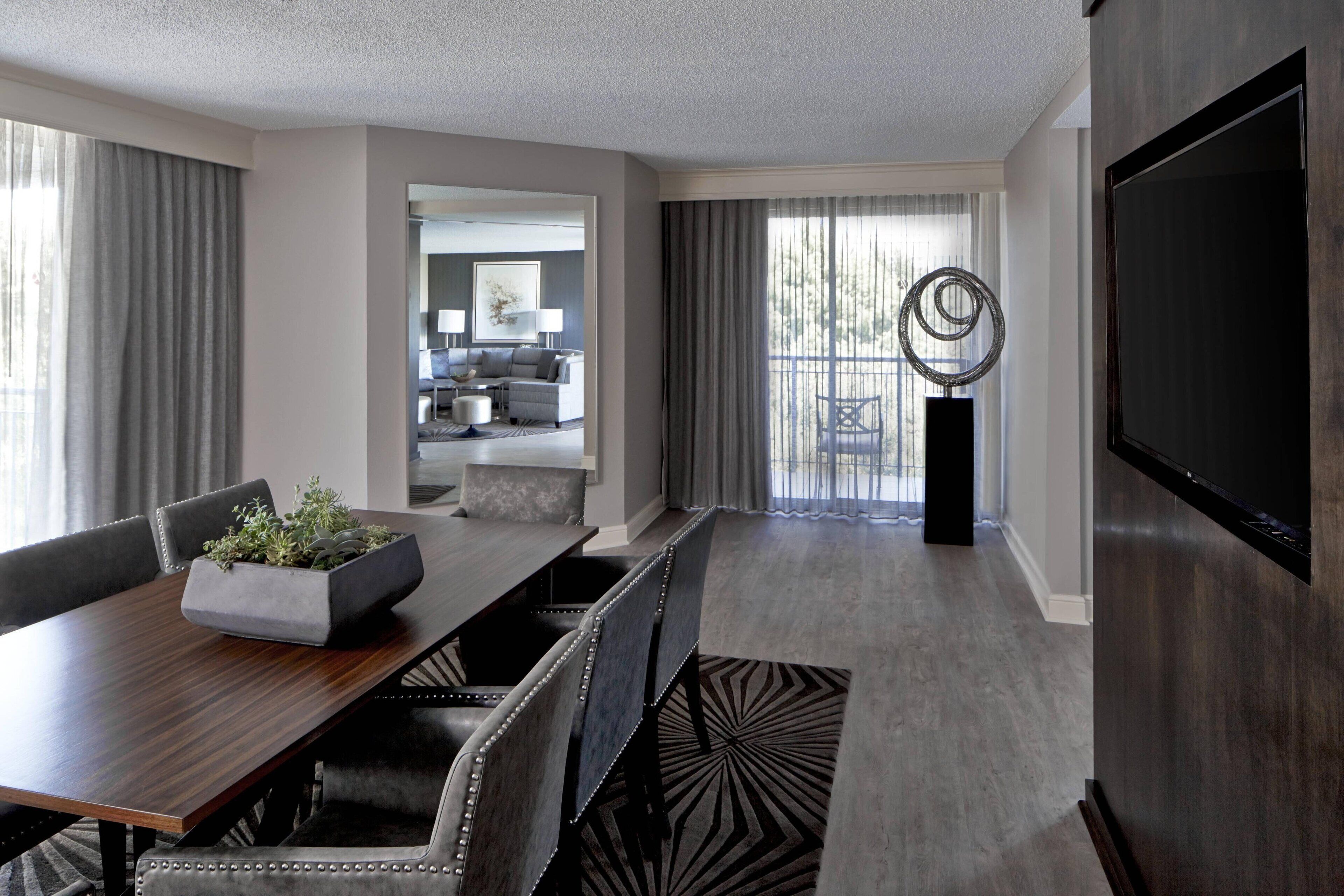 A modern hotel room with a dining area, a living room with a sofa and coffee table, and a large TV on the wall.