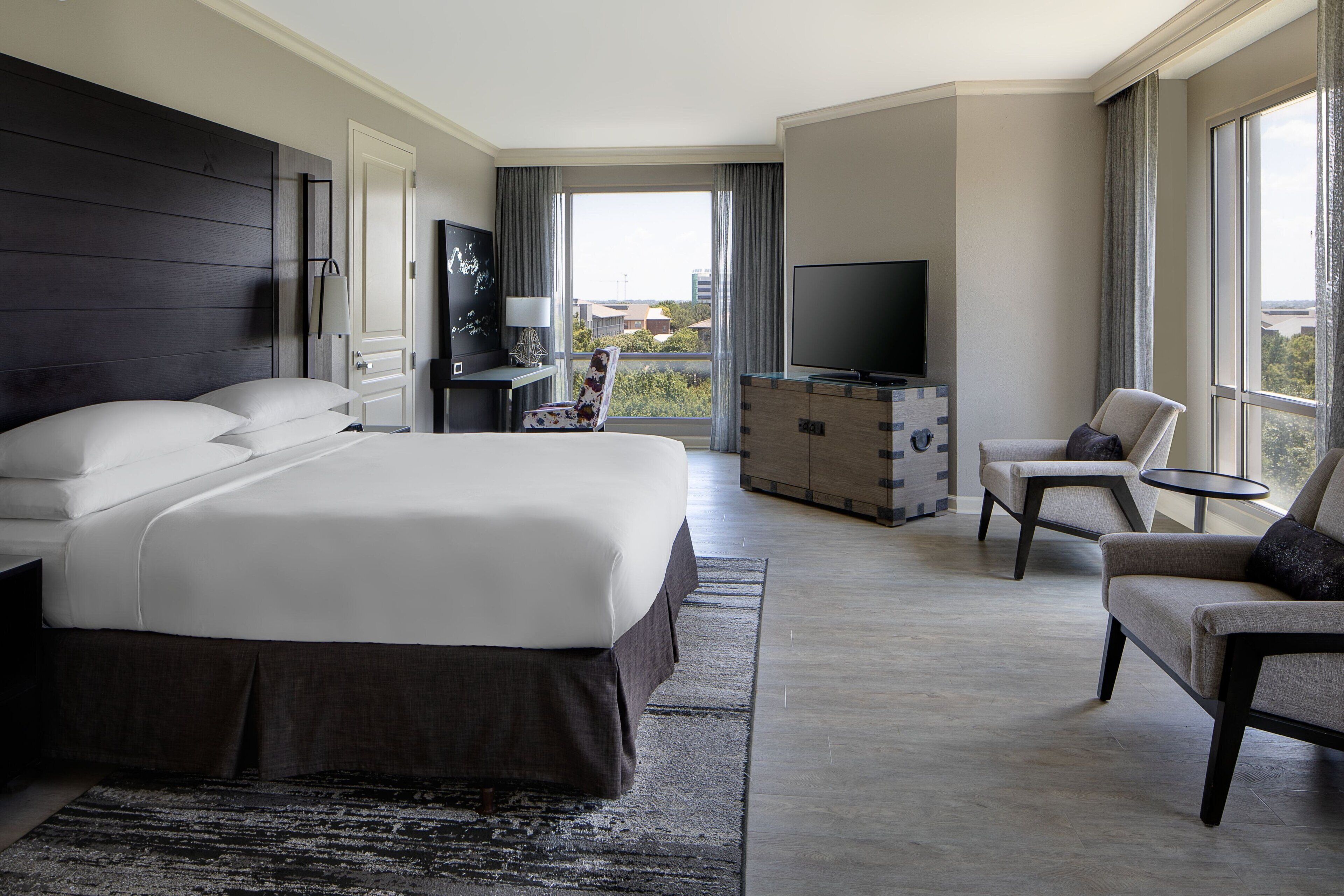 A modern hotel room with a large bed, a TV, a seating area with a chair and a small table, and a view of the city through large windows.