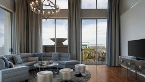 A modern living room with a sectional sofa, ottomans, a round coffee table, a TV on a cabinet, and large windows with a view of the city. - Dallas/Plano Marriott at Legacy Town Center (Plano)