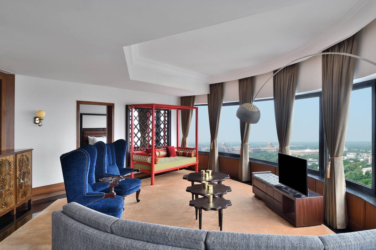 A modern hotel room with a large window, a sofa, a red daybed, a TV, and a small table.