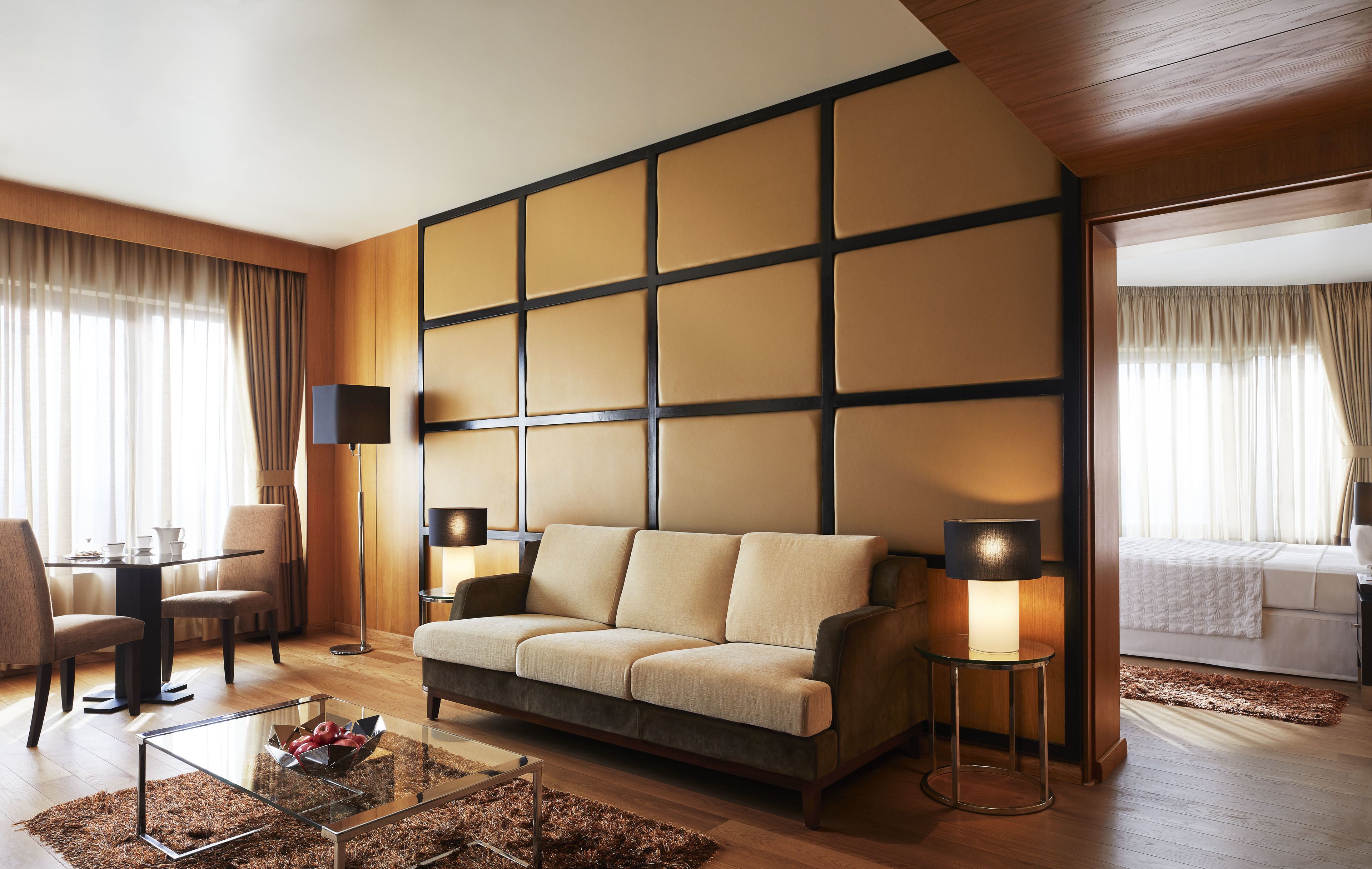 A modern hotel room with a sofa, two lamps, a glass coffee table, and a dining area with chairs and a table.