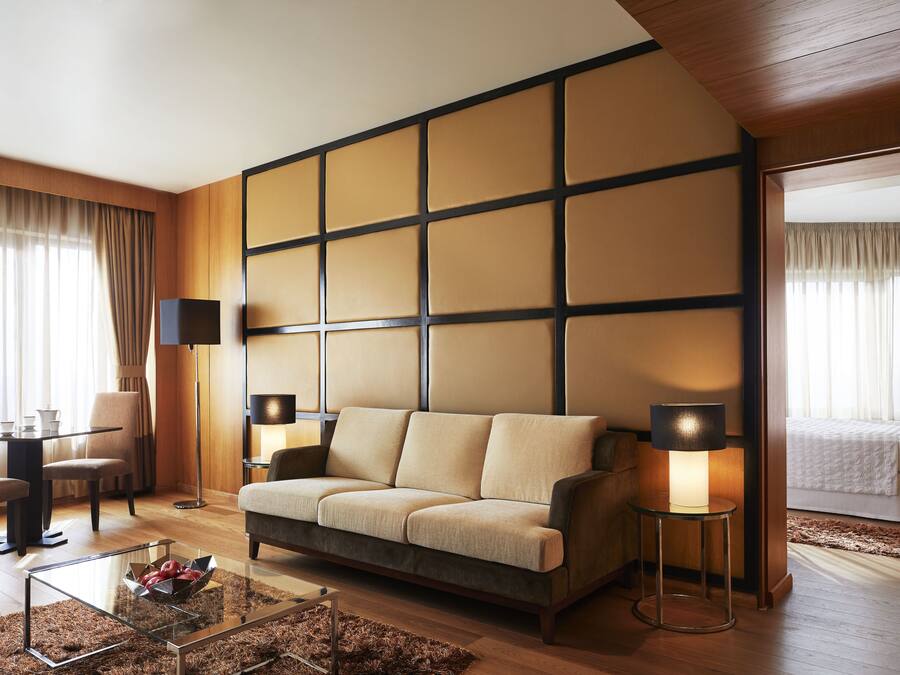 A modern hotel room with a sofa, two lamps, a glass coffee table, and a dining area with chairs and a table.