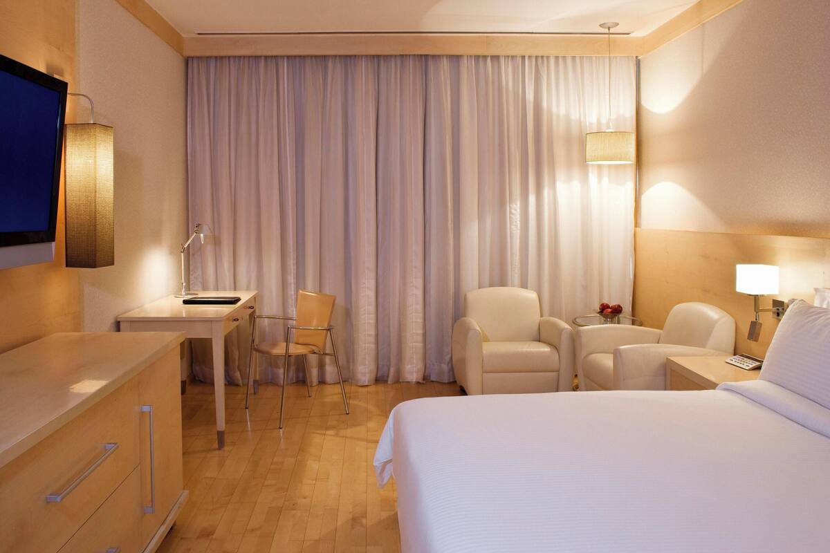 A hotel room with a bed, a desk, two chairs, a TV, and a window with curtains.