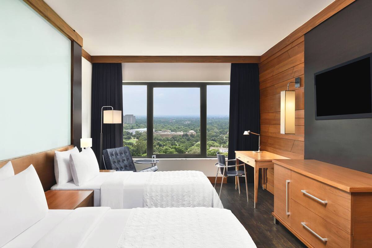 A hotel room with two beds, a desk, a chair, and a large window offering a city view.