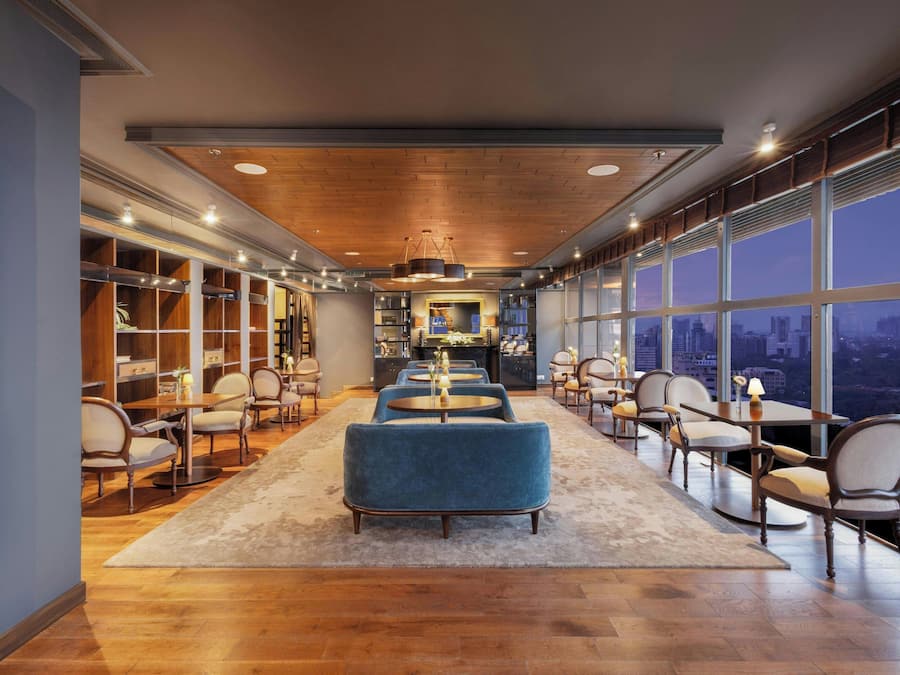 A spacious hotel lobby with a long wooden table, comfortable seating, and large windows offering a city view.