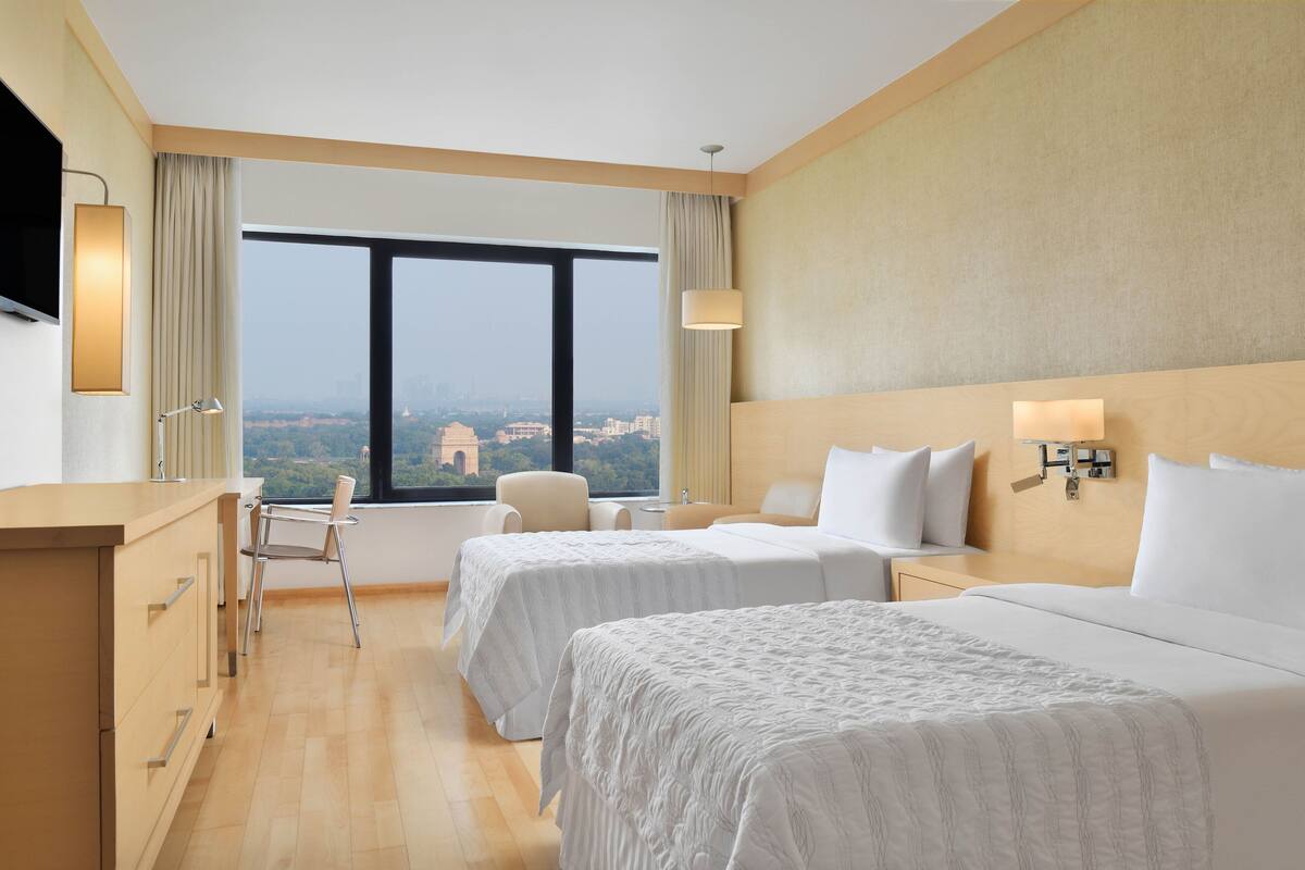 A hotel room with two beds, a desk, a chair, a TV, and a large window offering a city view.