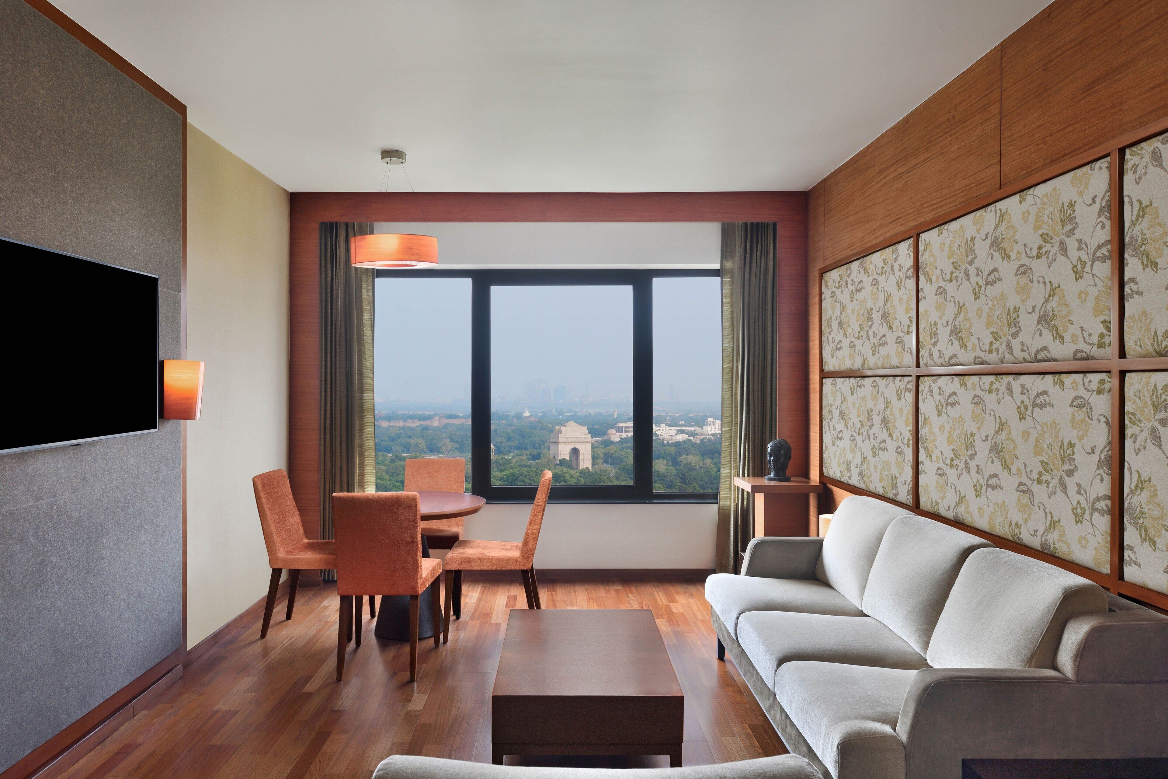 A modern hotel room with a large window, a flat-screen TV, a sofa, a dining table with chairs, and a decorative panel.