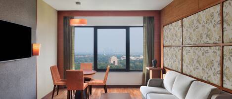 Habitación de hotel moderna con un ventanal grande, un televisor de pantalla plana, un sofá, una mesa de comedor con sillas y un panel decorativo.