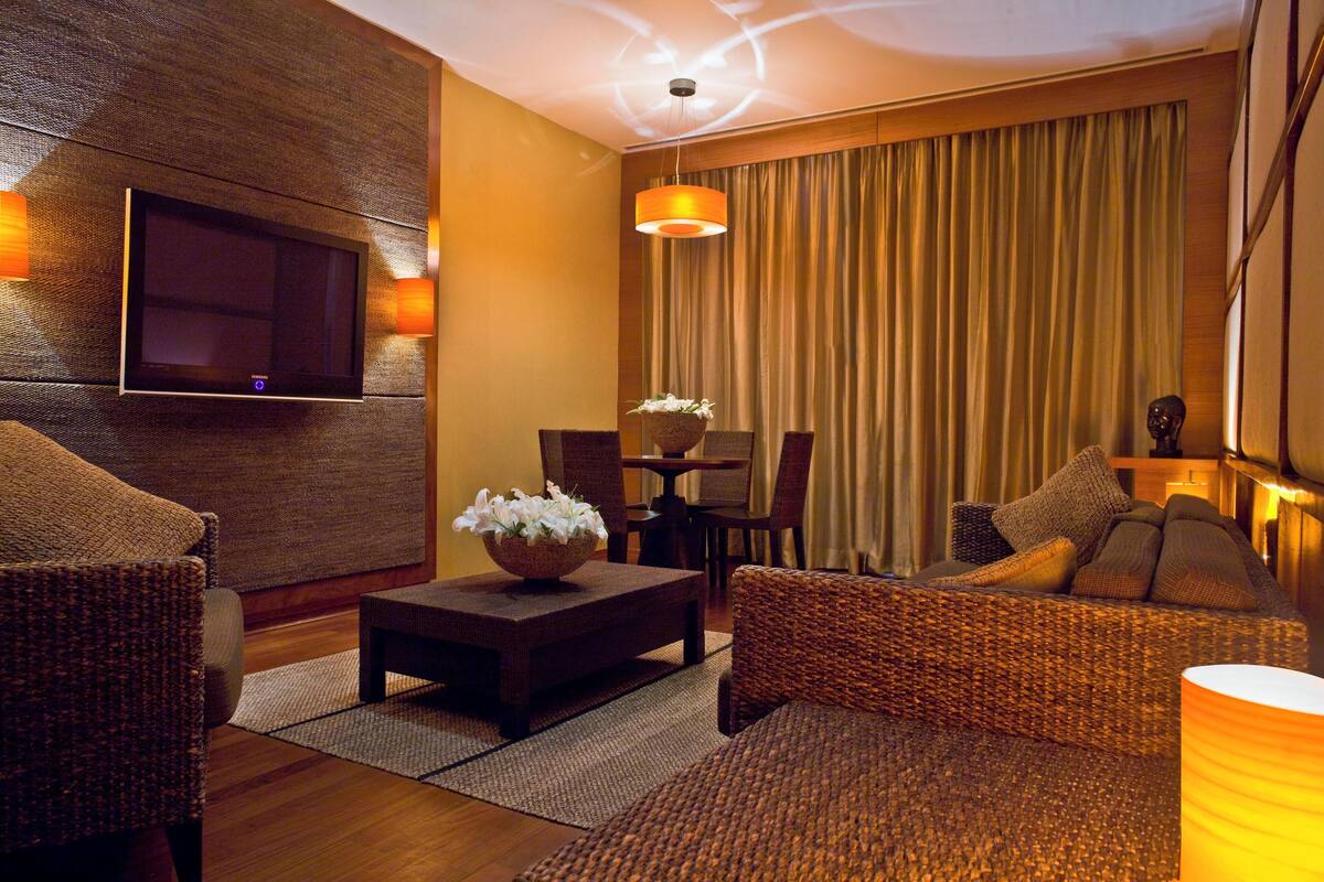 A modern living room with a flat-screen TV mounted on a textured wall, a wicker sofa, a small coffee table with a floral centerpiece, and a dining area with a table and chairs.