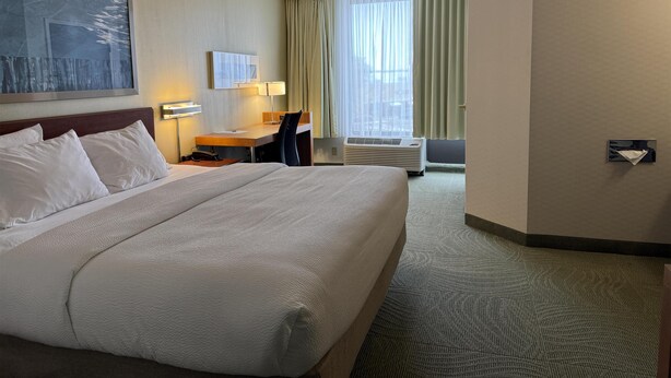 Ein Hotelzimmer mit einem großen Bett, einem Schreibtisch mit Stuhl, einer Schreibtischlampe, einem Fenster mit Vorhängen und einer wandmontierten Klimaanlage.