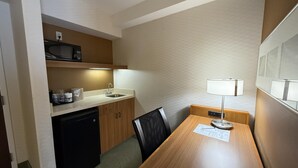 Ein kompaktes Hotelzimmer mit Küchenzeile, einem Holzesstisch, einem schwarzen Stuhl, einem kleinen Schreibtisch mit Lampe und einem am Wand montierten Fernseher.