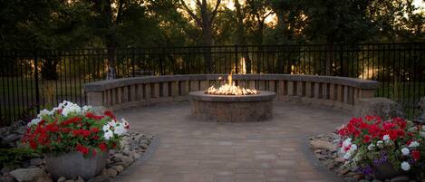 A circular stone fire pit surrounded by a stone pathway, with flower beds on either side.