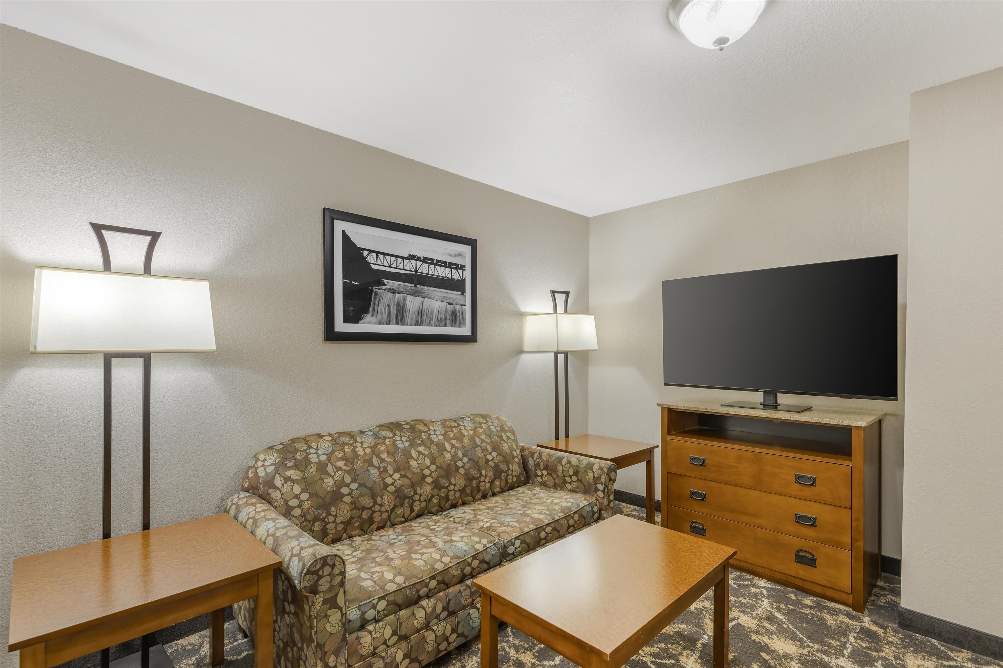 Habitación de hotel con un sofá, dos mesas, un televisor sobre un soporte de madera y dos lámparas de pie.
