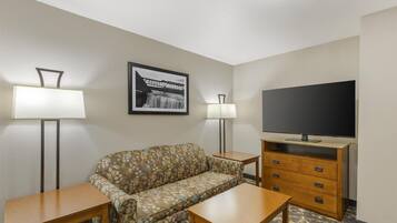 Habitación de hotel con un sofá, dos mesas, un televisor sobre un soporte de madera y dos lámparas de pie.