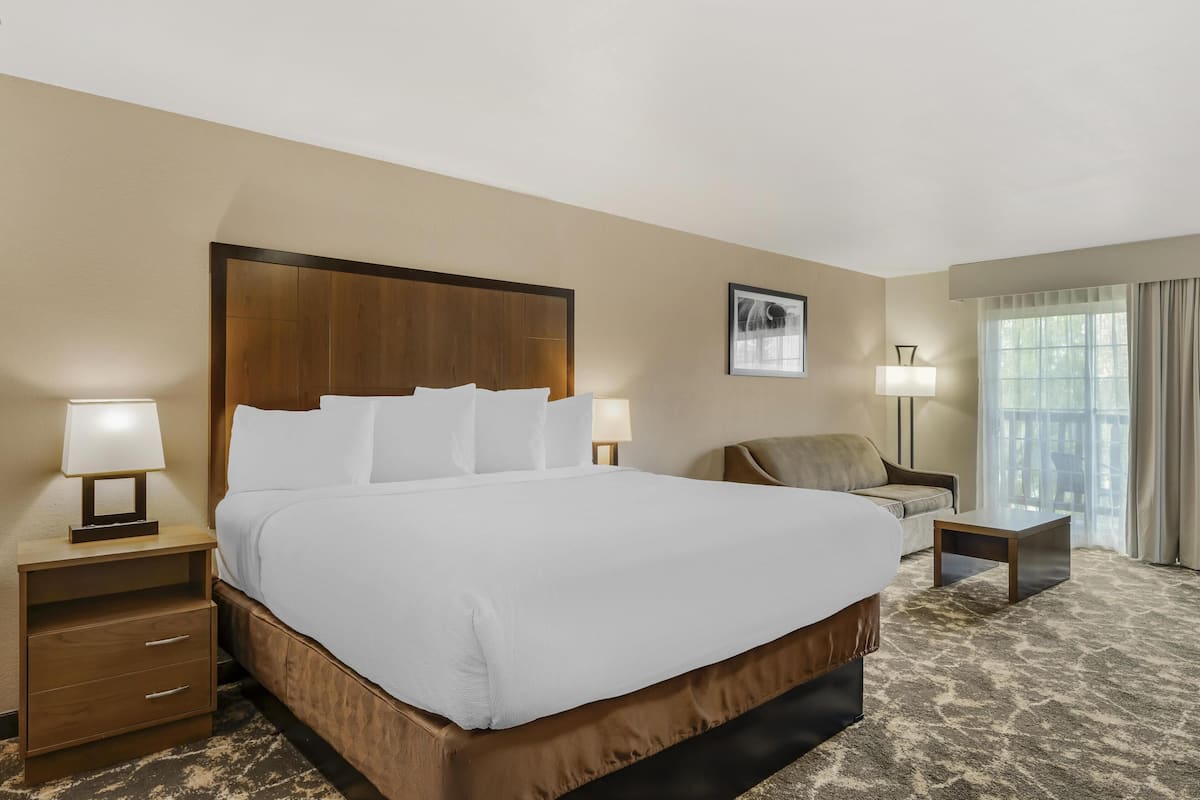 Ein Hotelzimmer mit einem großen Bett, zwei Nachttischen mit Lampen, einem Sessel, einem kleinen Tisch und einem Fenster mit Vorhängen.