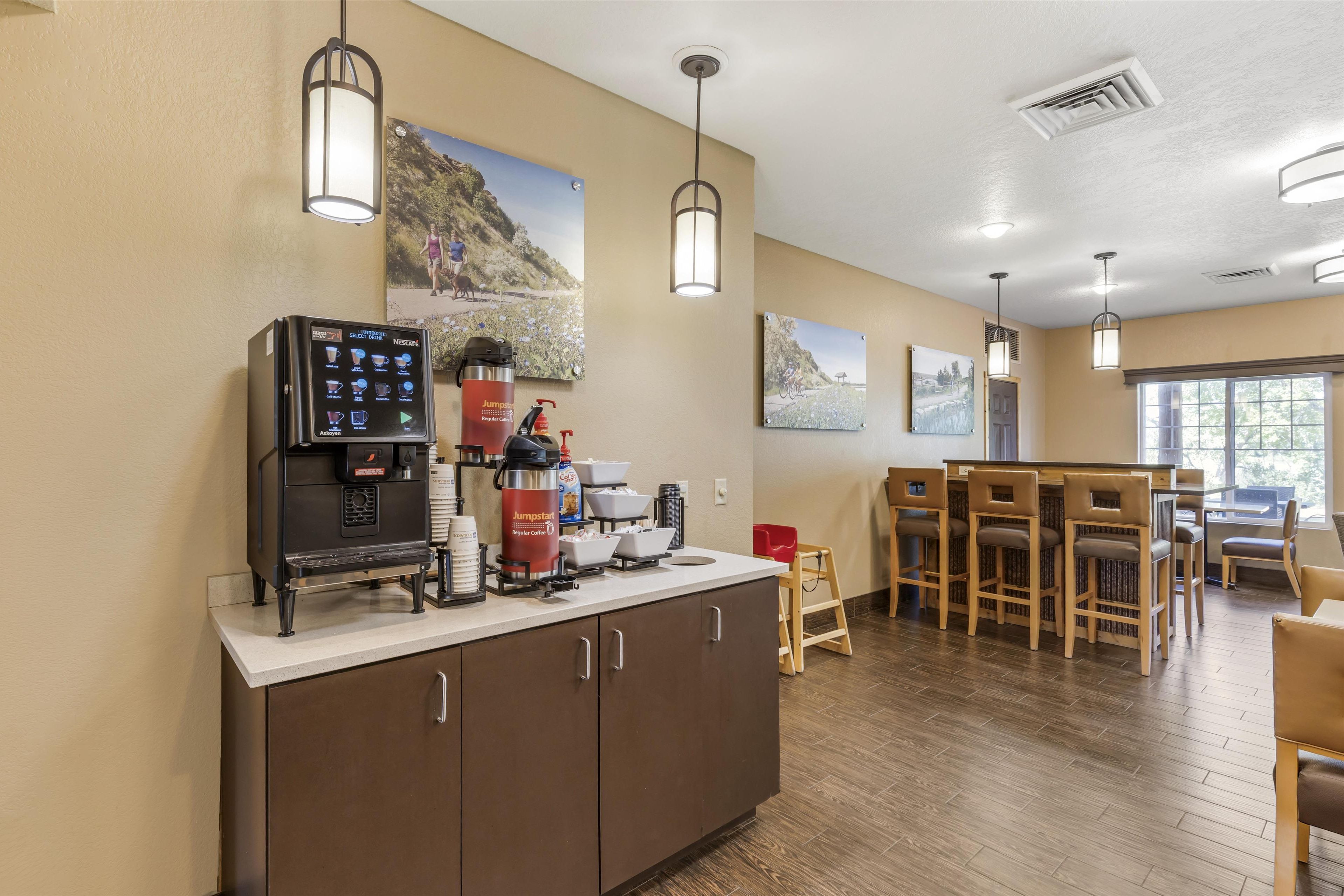A kitchen area with a coffee machine, a red coffee dispenser, and a microwave. There are wooden chairs and a table in the background.