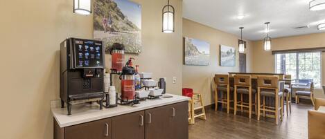 A kitchen area with a coffee machine, a red coffee dispenser, and a microwave. There are wooden chairs and a table in the background.