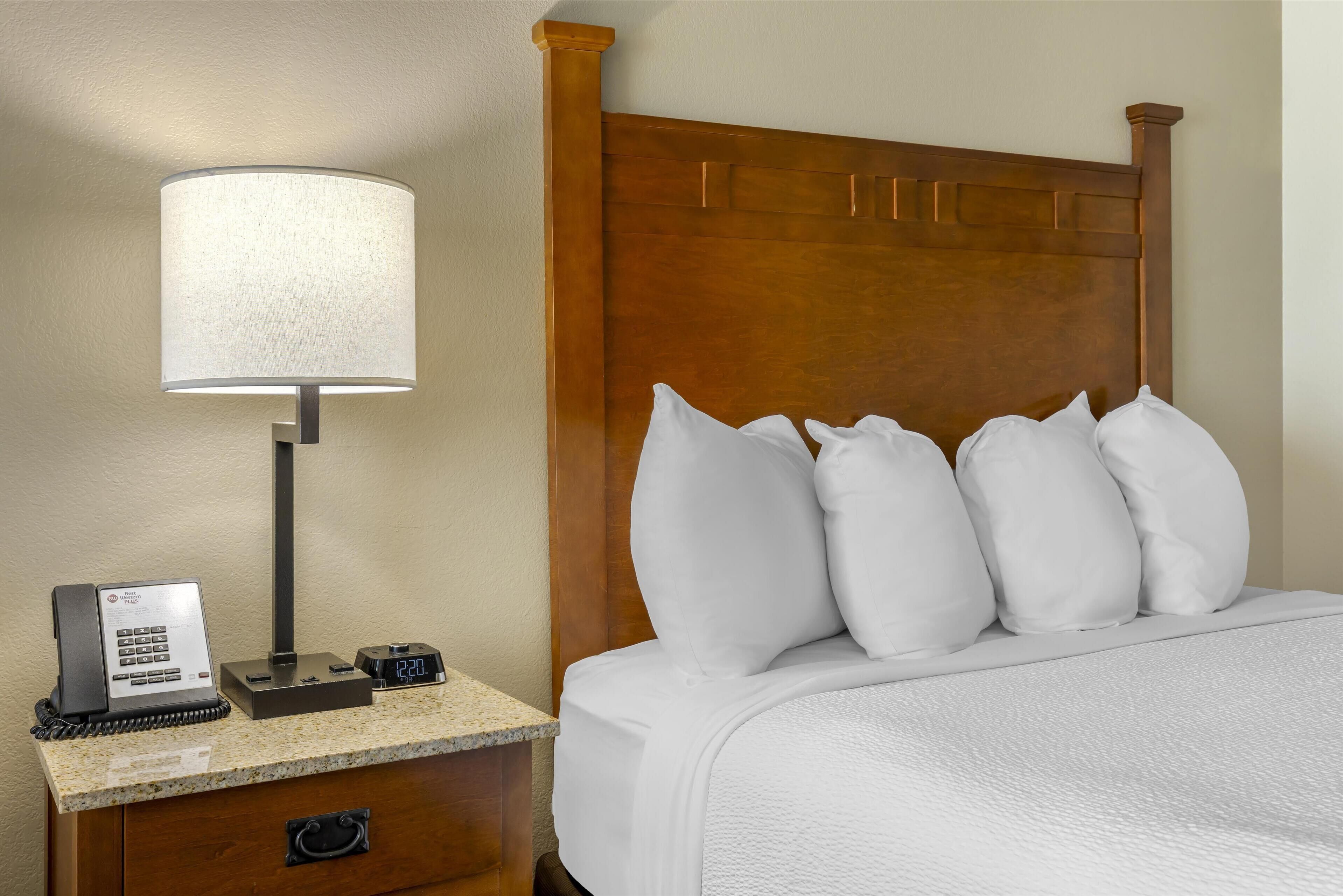A hotel room with a wooden headboard, a bedside table with a lamp, a clock, and a telephone.