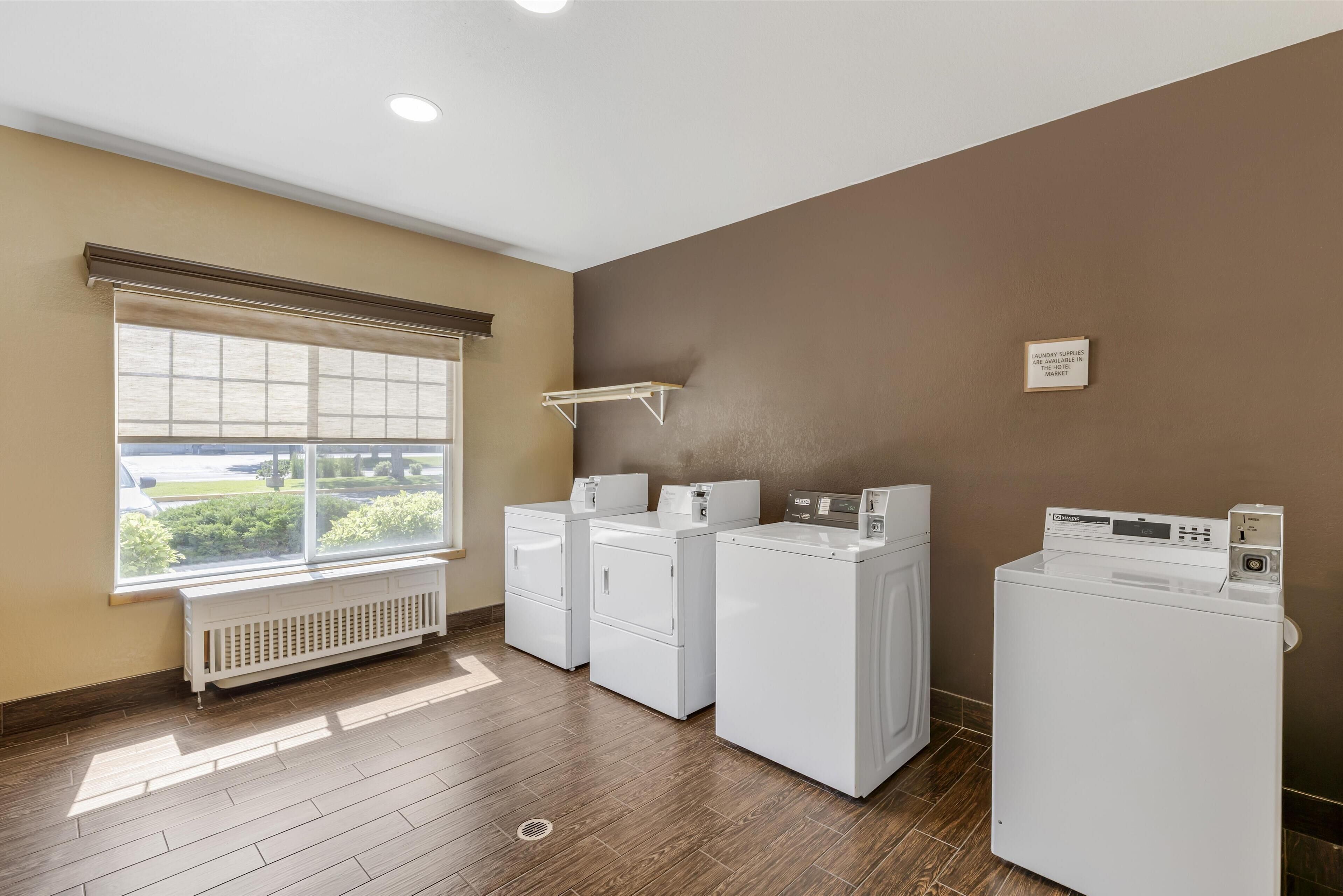 A laundry room with three stacked washers and dryers, a window with blinds, and a radiator.