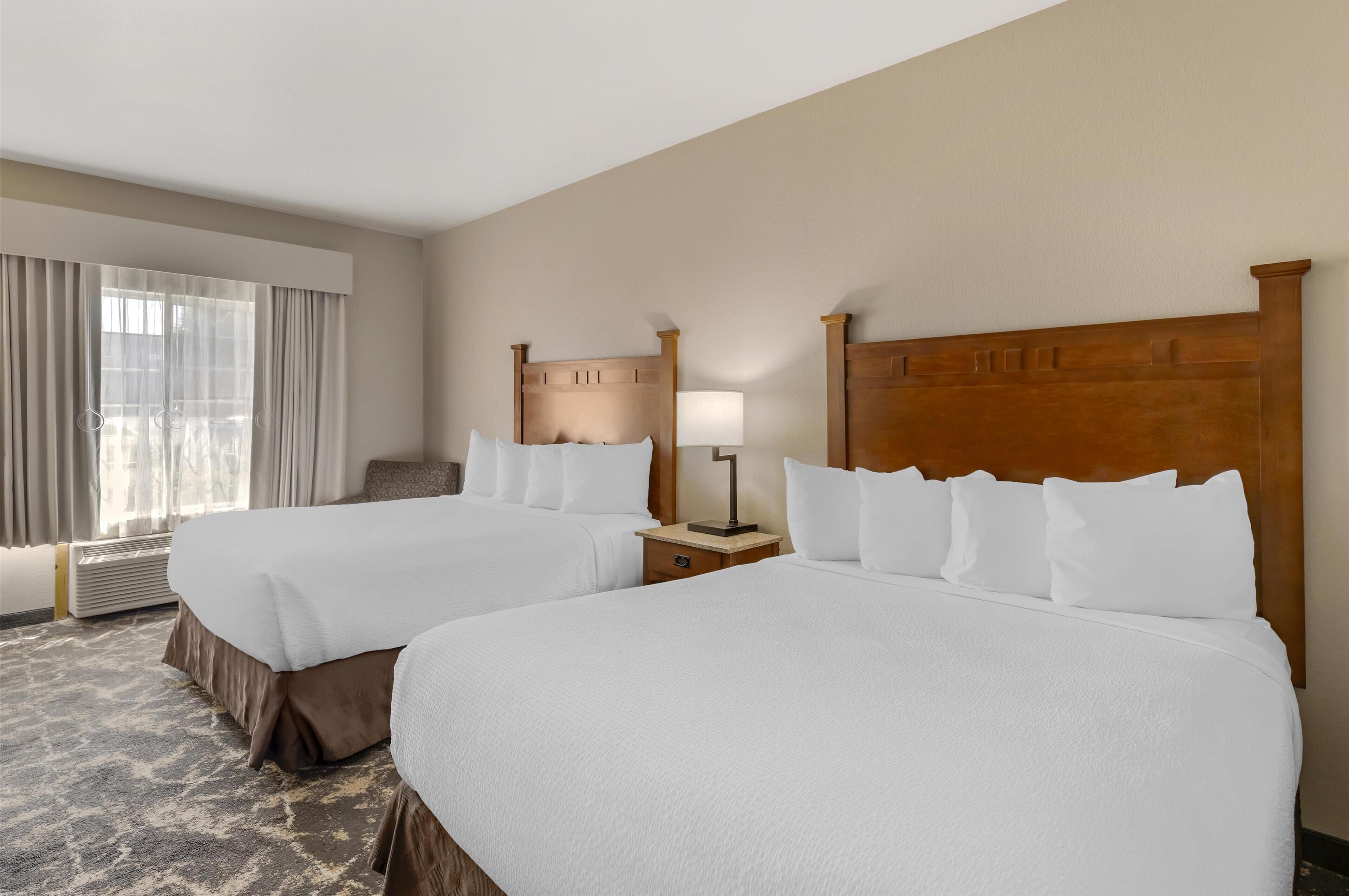 A hotel room with two beds, a nightstand with a lamp, a window with curtains, and a carpeted floor.