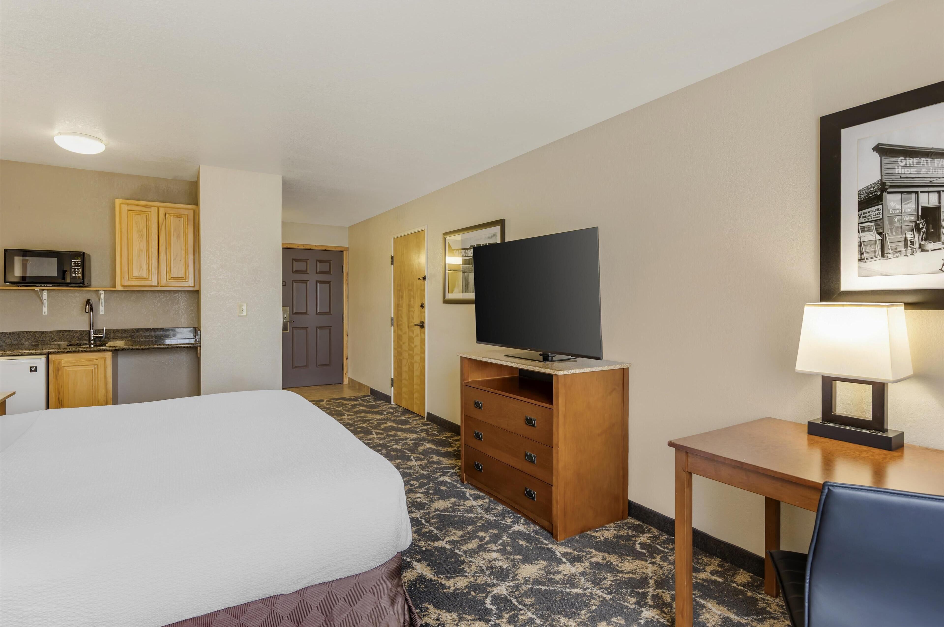 A hotel room with a bed, a television on a wooden stand, a desk with a lamp, and a kitchenette with a microwave and cabinets.
