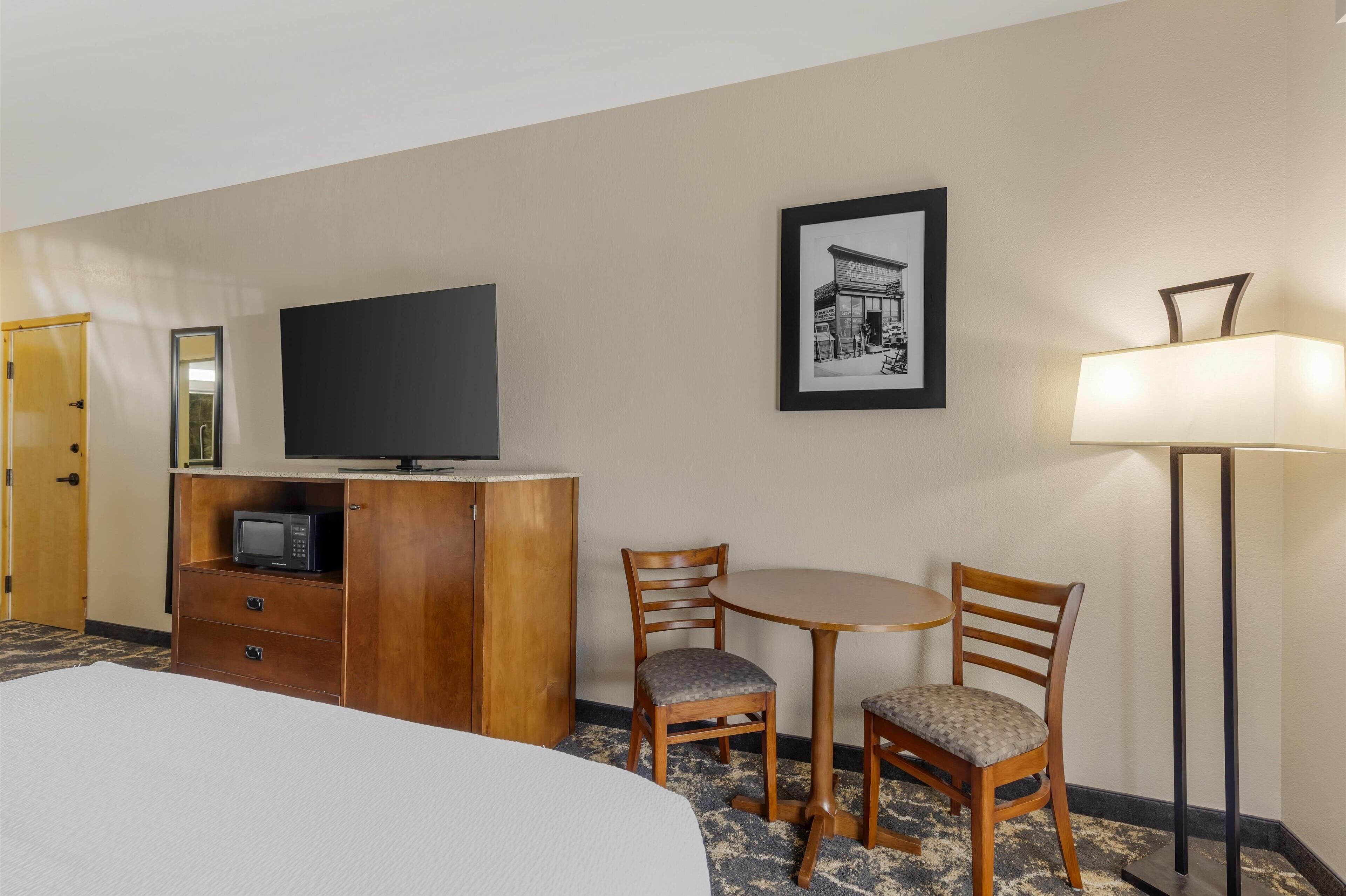 Une chambre d’hôtel avec un lit, un téléviseur posé sur une commode en bois, une petite table avec deux chaises, un tableau encadré au mur et un lampadaire.