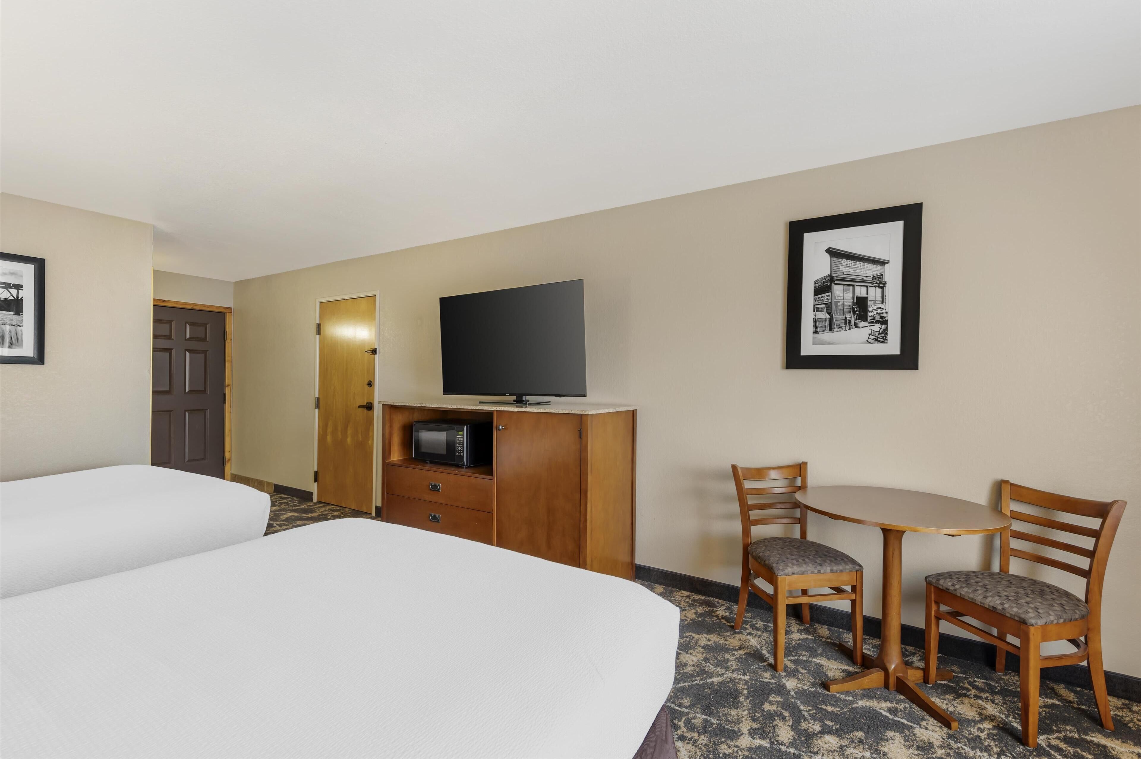 A hotel room with two beds, a wooden desk with a chair, a television, and framed pictures on the wall.